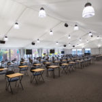 Interior of a large temporary building set up as an exam hall with rows of desks and lighting.