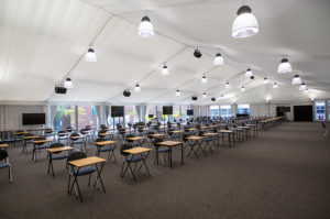 Interior of a large temporary building set up as an exam hall with rows of desks and lighting.